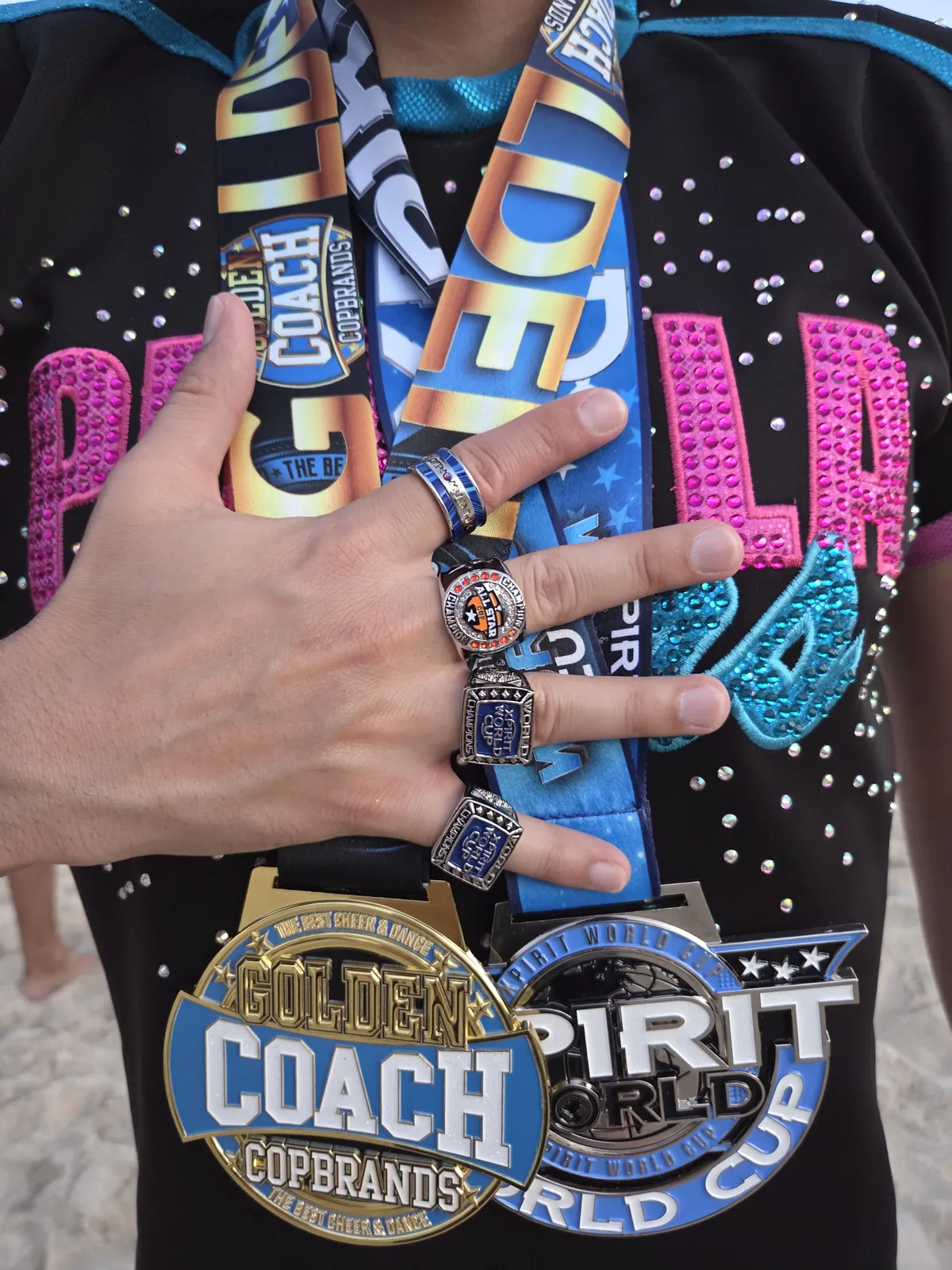 A hand wearing championship rings over cheerleading medal ribbons, including a COPBRANDS Golden Coach medal and a Spirit World Cup medal.