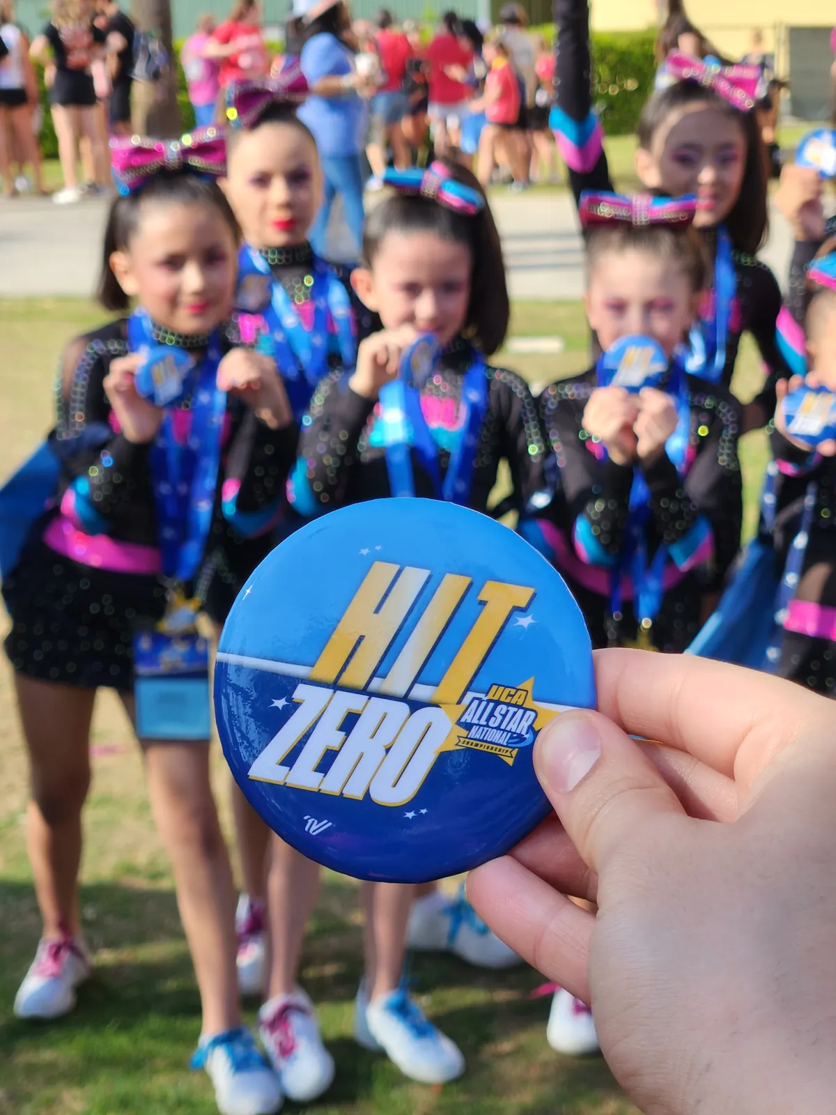 A youth cheerleading team in uniform and bows shows their medals; in front, a hand holds a “Hit Zero” UCA All Star Nationals button.